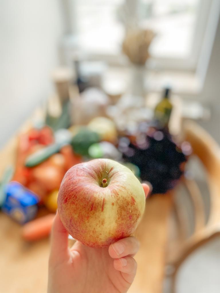 An apple being held up in front of a table of fresh groceries. 